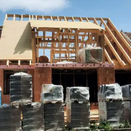 Budowa domu murowanego: widok na konstrukcję dachu w trakcie montażu, z paletami materiałów budowlanych przy chodniku i fragmentem starego dachu po prawej.