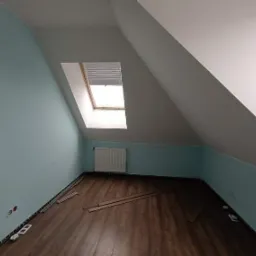 Poddasze po remoncie: świeżo pomalowane ściany w odcieniach błękitu i szarości, nowa podłoga z paneli, widoczne listwy przypodłogowe i fragmenty paneli na podłodze przed montażem, okno dachowe...