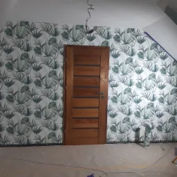 Ściana z tapetą w motywy roślinne, brązowe drzwi z poziomymi panelami, widoczne przewody elektryczne zwisające z sufitu, podłoga zabezpieczona folią malarską.