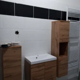 Montaż mebli łazienkowych z drewnopodobnymi frontami na tle białych płytek z czarnym pasem dekoracyjnym; widoczne wystające kable elektryczne.