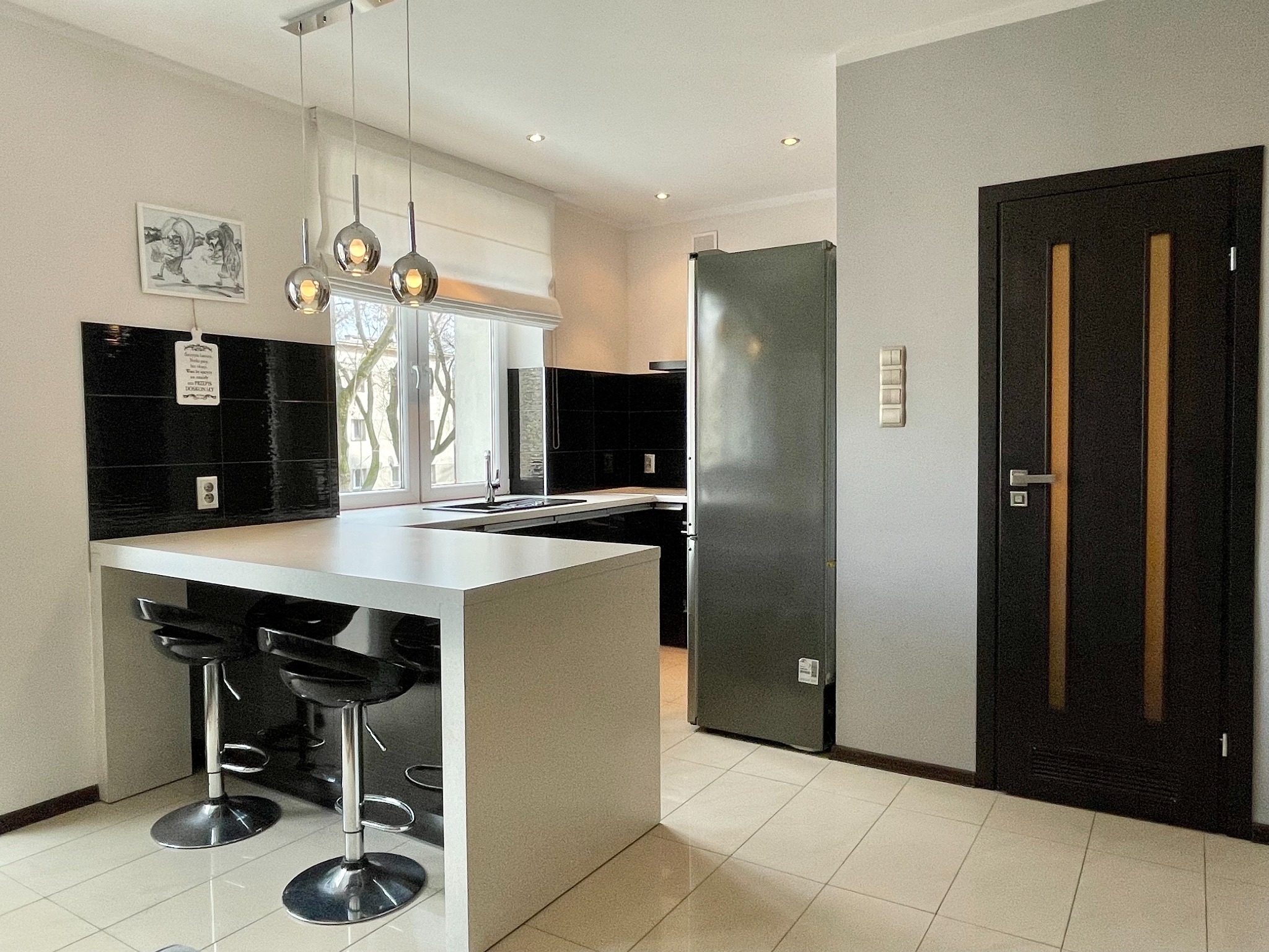 Nowoczesna kuchnia z wyspą i czarnymi hokerami, czarne płytki na ścianie, okno z roletą rzymską, srebrna lodówka i ciemne drzwi z jasnymi wstawkami.