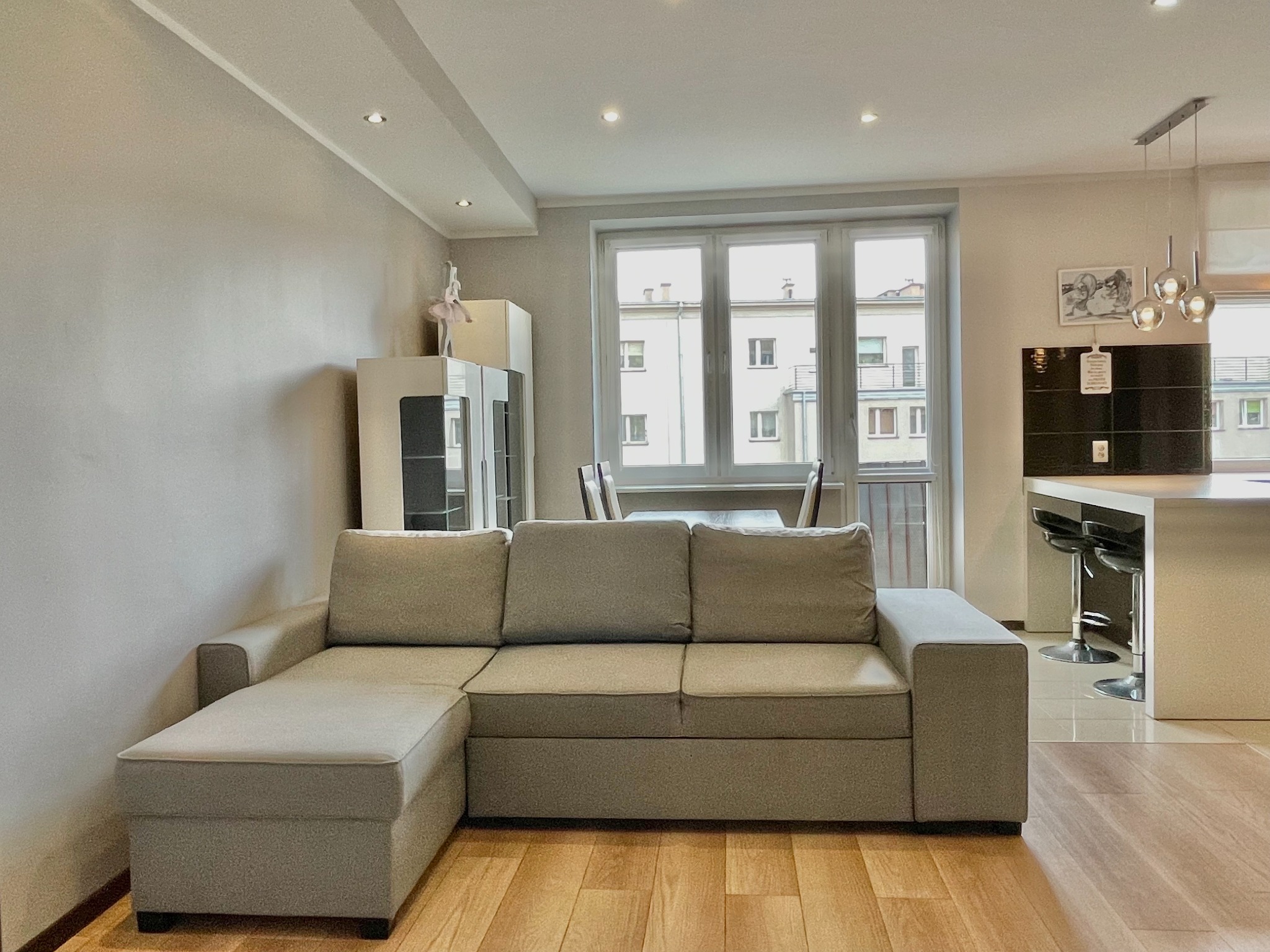 Jasne, przestronne wnętrze mieszkania z szarą sofą narożną, drewnianą podłogą i widokiem na budynki zza okna.