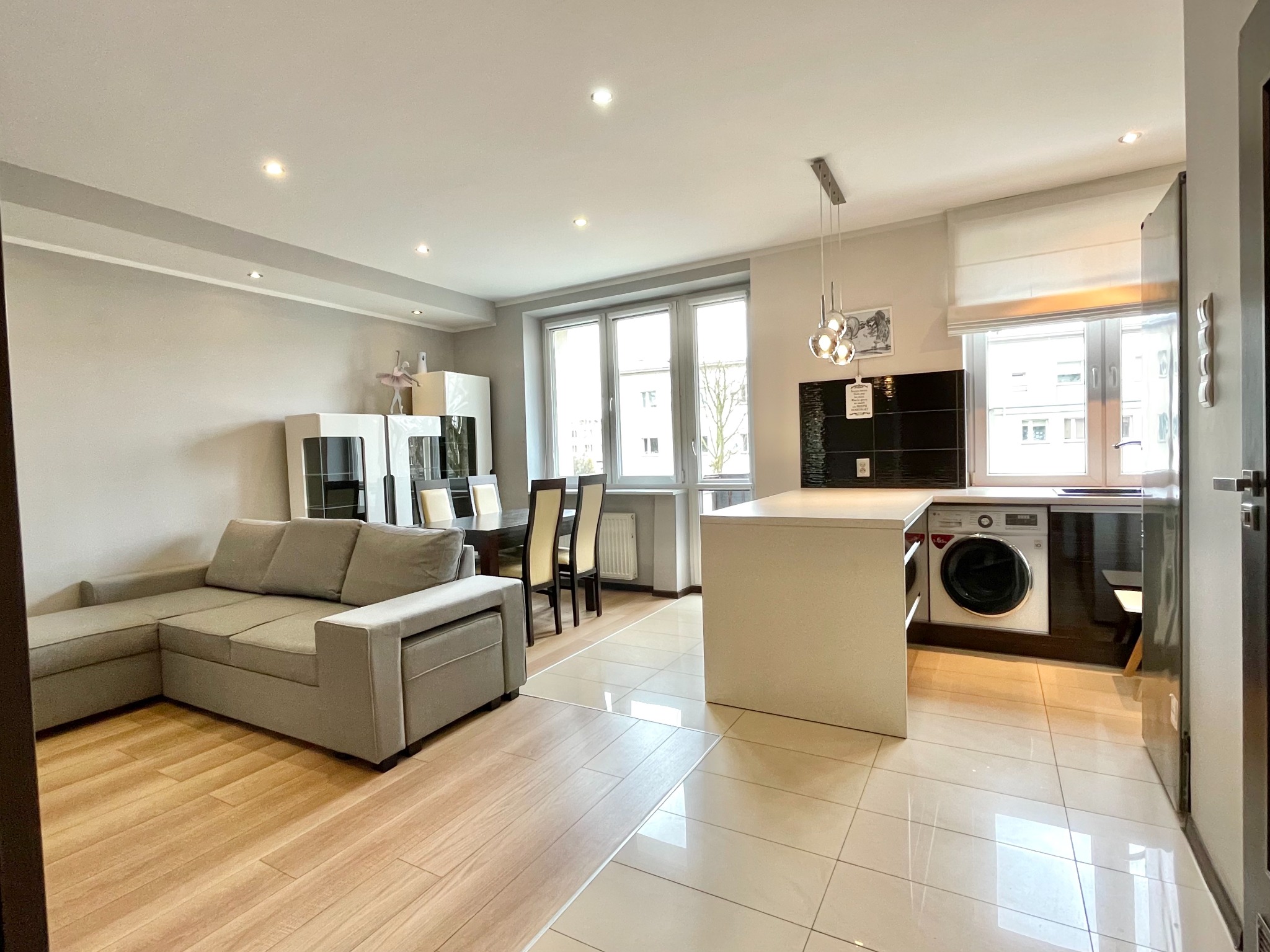 Jasne, przestronne wnętrze mieszkania z narożną sofą, stołem z krzesłami i otwartą kuchnią z pralką w zabudowie.