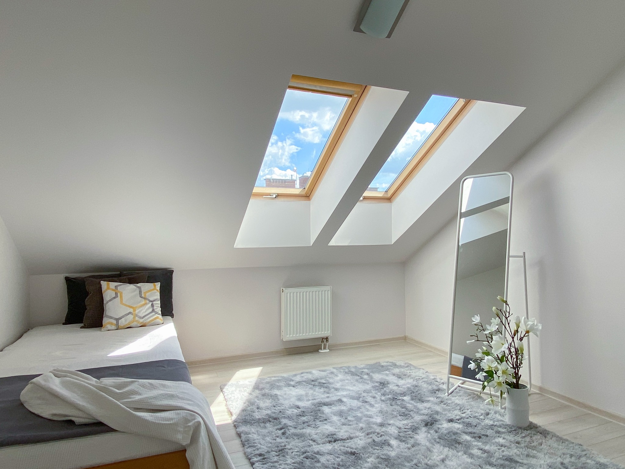 Jasne poddasze z łóżkiem, puszystym dywanem, stojącym lustrem i dwoma oknami dachowymi z widokiem na błękitne niebo.