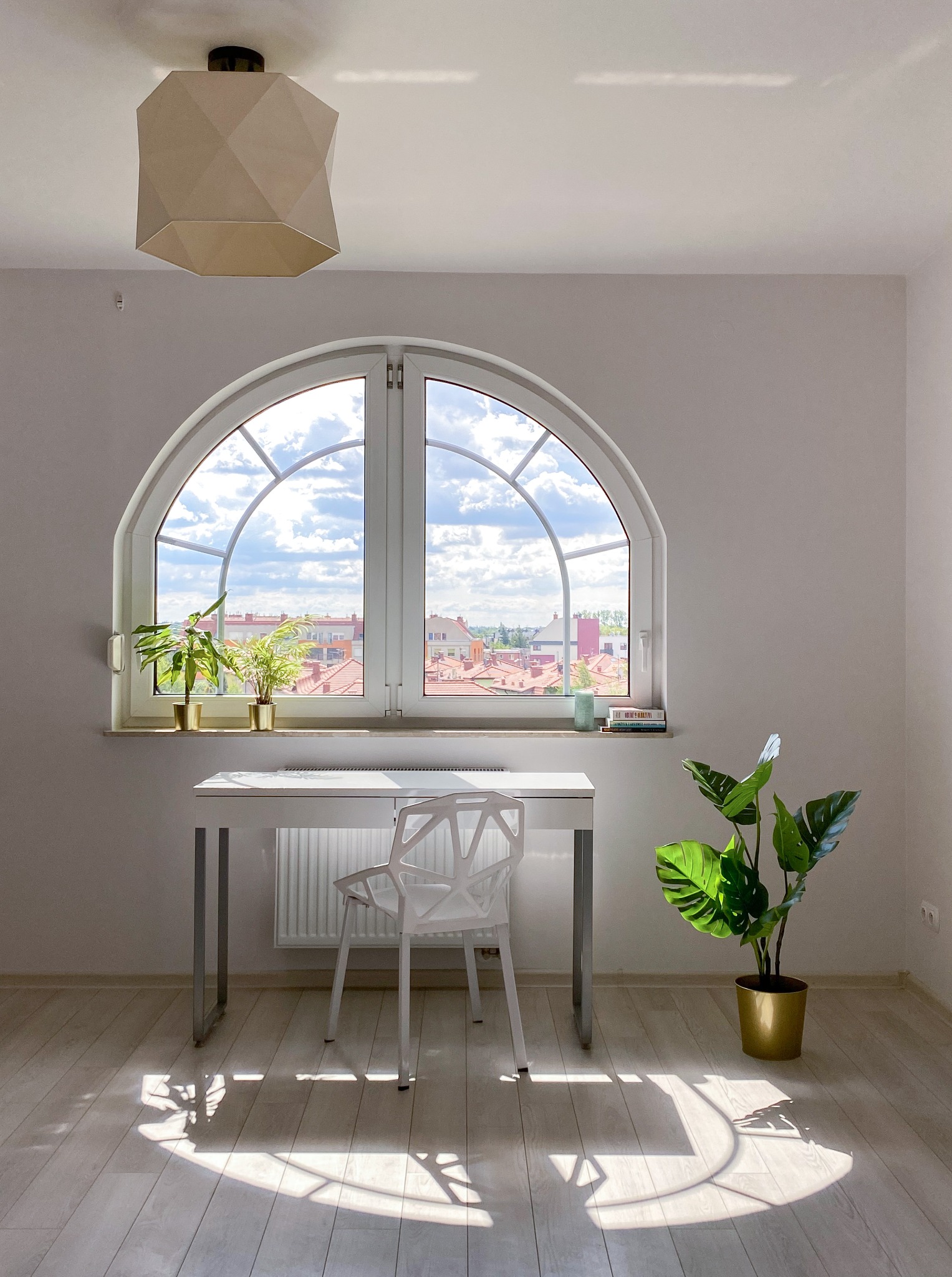 Jasne wnętrze z biurkiem, krzesłem i dużym oknem łukowym z widokiem na miasto, roślina doniczkowa w złotym wazonie i geometryczna lampa wisząca.