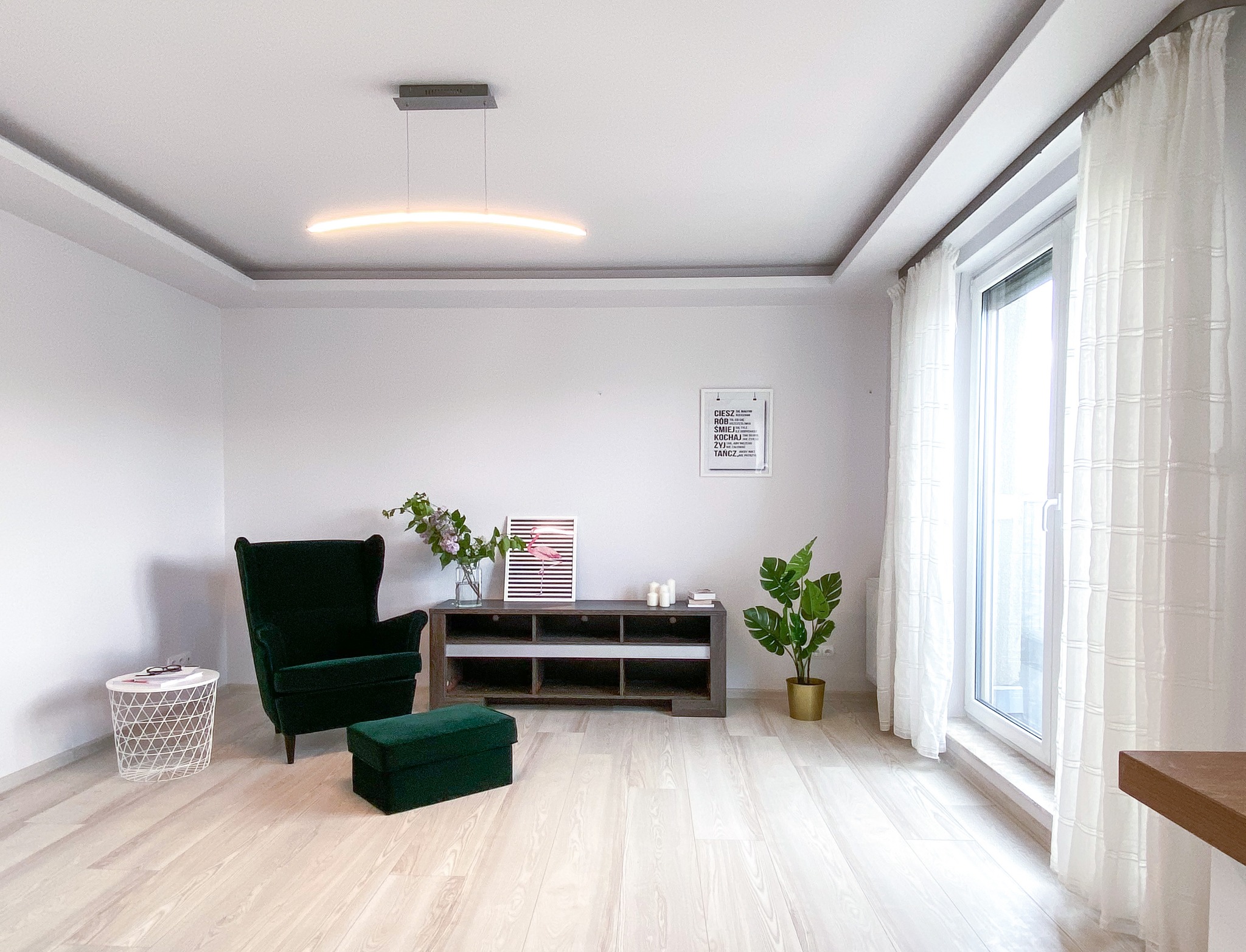 Jasne, minimalistyczne wnętrze: zielony fotel z podnóżkiem, drewniana komoda z dekoracjami, roślina w złotej doniczce, okno z białymi zasłonami i nowoczesna lampa sufitowa.