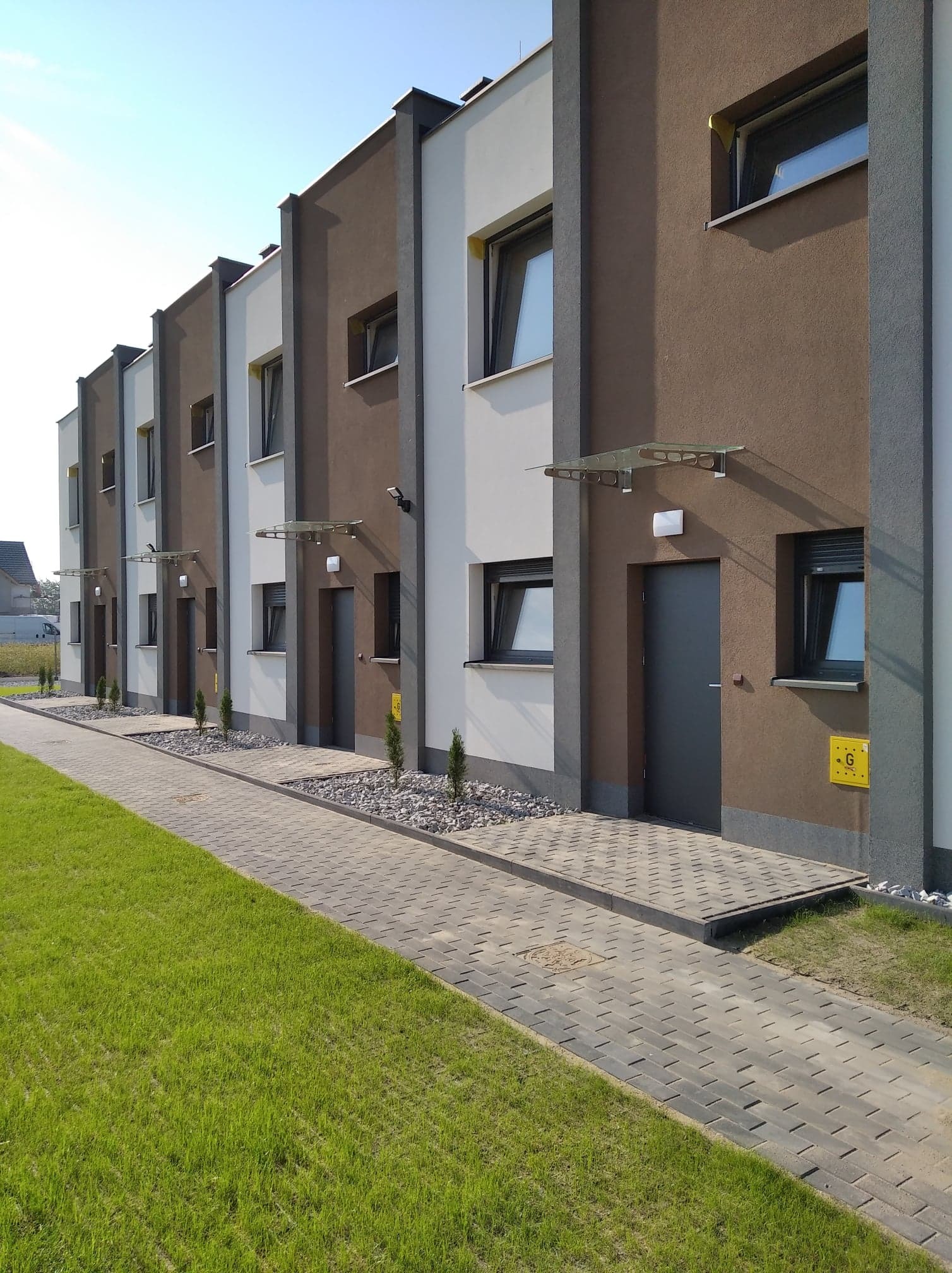 Rząd nowo wybudowanych, dwukondygnacyjnych domów szeregowych z brązowymi i białymi elewacjami, szarymi drzwiami wejściowymi i szklanymi daszkami, widok z poziomu trawnika i chodnika z kostki brukowej.