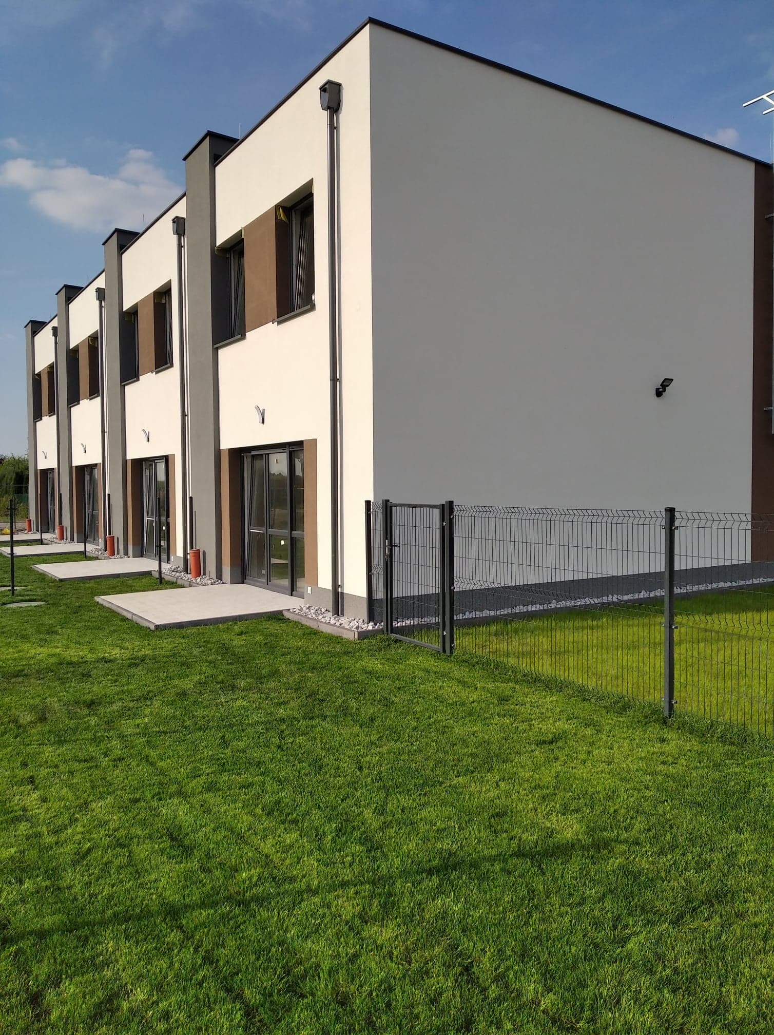 Szereg nowo wybudowanych, dwukondygnacyjnych domów w zabudowie szeregowej z ogrodami, widok z zewnątrz przez metalowe ogrodzenie.