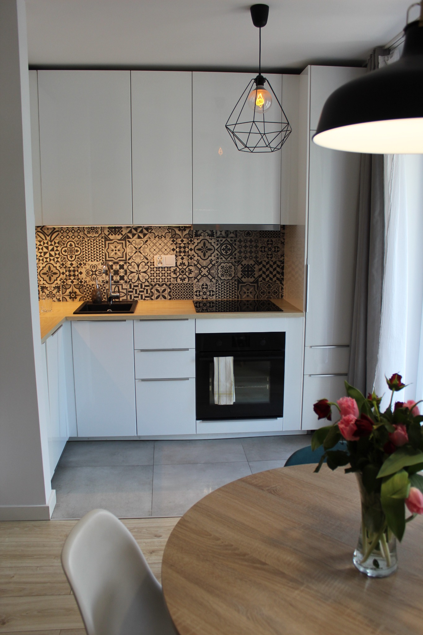 Nowoczesna kuchnia z białymi szafkami, czarną armaturą i dekoracyjnymi płytkami backsplash w geometryczne wzory, oświetlona lampą wiszącą z żarówką Edison, z bukietem róż na drewnianym stole...