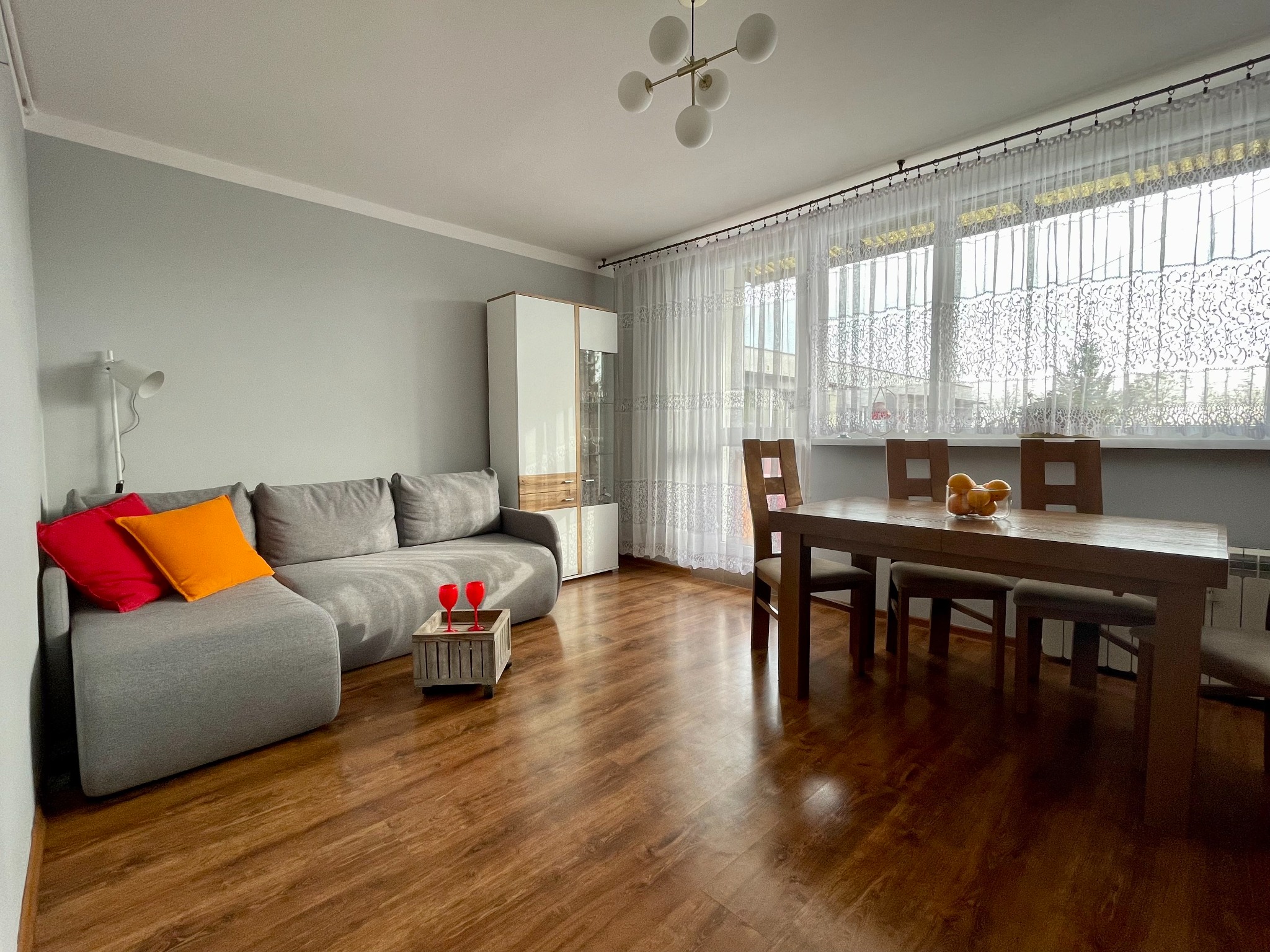 Jasnoszary salon z drewnianą podłogą, szarą sofą z pomarańczową i czerwoną poduszką, drewnianym stołem z owocami i białymi zasłonami.