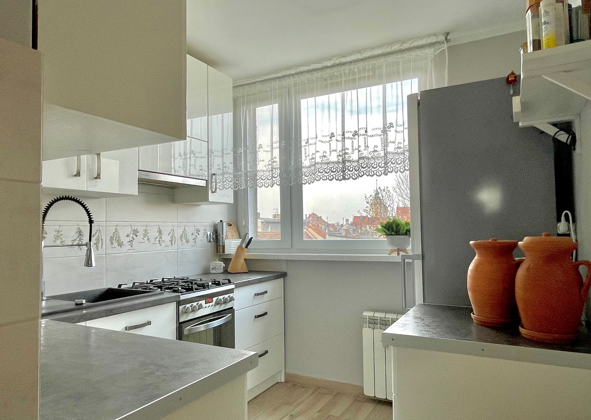 Jasna kuchnia z białymi szafkami, szarym blatem i oknem z firanką z motywem kawy, widok na miasto w tle, dwa gliniane dzbanki na blacie.