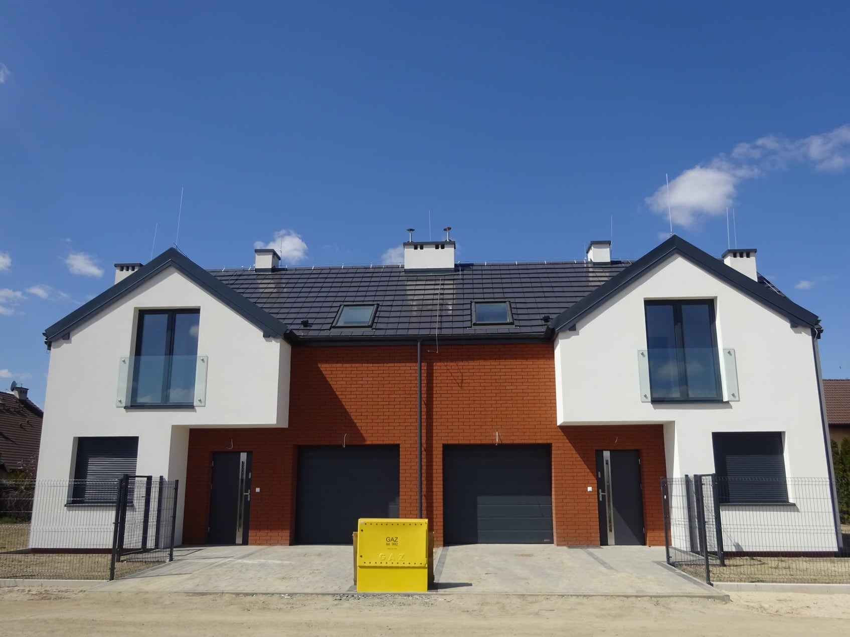 Dwa bliźniacze domy w nowoczesnym stylu z białymi ścianami i elementami z czerwonej cegły, grafitowe dachy i okna, żółta skrzynka gazowa na pierwszym planie, błękitne niebo w tle.