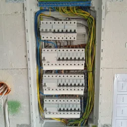 Otwarta skrzynka elektryczna z zamontowanymi bezpiecznikami i podłączonymi przewodami w kolorach niebieskim, żółtym i zielonym, zamocowana na surowej, otynkowanej ścianie. Widoczny fragment...