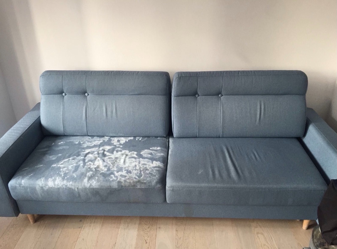 Szara kanapa z widoczną plamą na jednym siedzisku, na drewnianej podłodze, przed czystą ścianą.