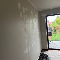 NORBUD Norbert Grzesiak - Ściana w trakcie szpachlowania z widocznymi łatami gładzi, zabezpieczona podłoga folią i osoba w tle, stojąca w otwartych drzwiach na zewnątrz.
