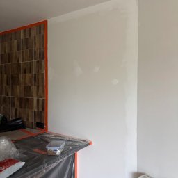 NORBUD Norbert Grzesiak - Ściana po szpachlowaniu, przygotowana do malowania. Widoczne zabezpieczenia folią malarską i taśmą. Fragment ściany z brązowymi płytkami.