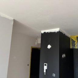 NORBUD Norbert Grzesiak - Wnętrze w trakcie malowania: czarna ściana z widocznymi śladami szpachlowania i świeżo pomalowany sufit na biało. Zabezpieczone folią drzwi i fragment ściany w odcieniach szarości.