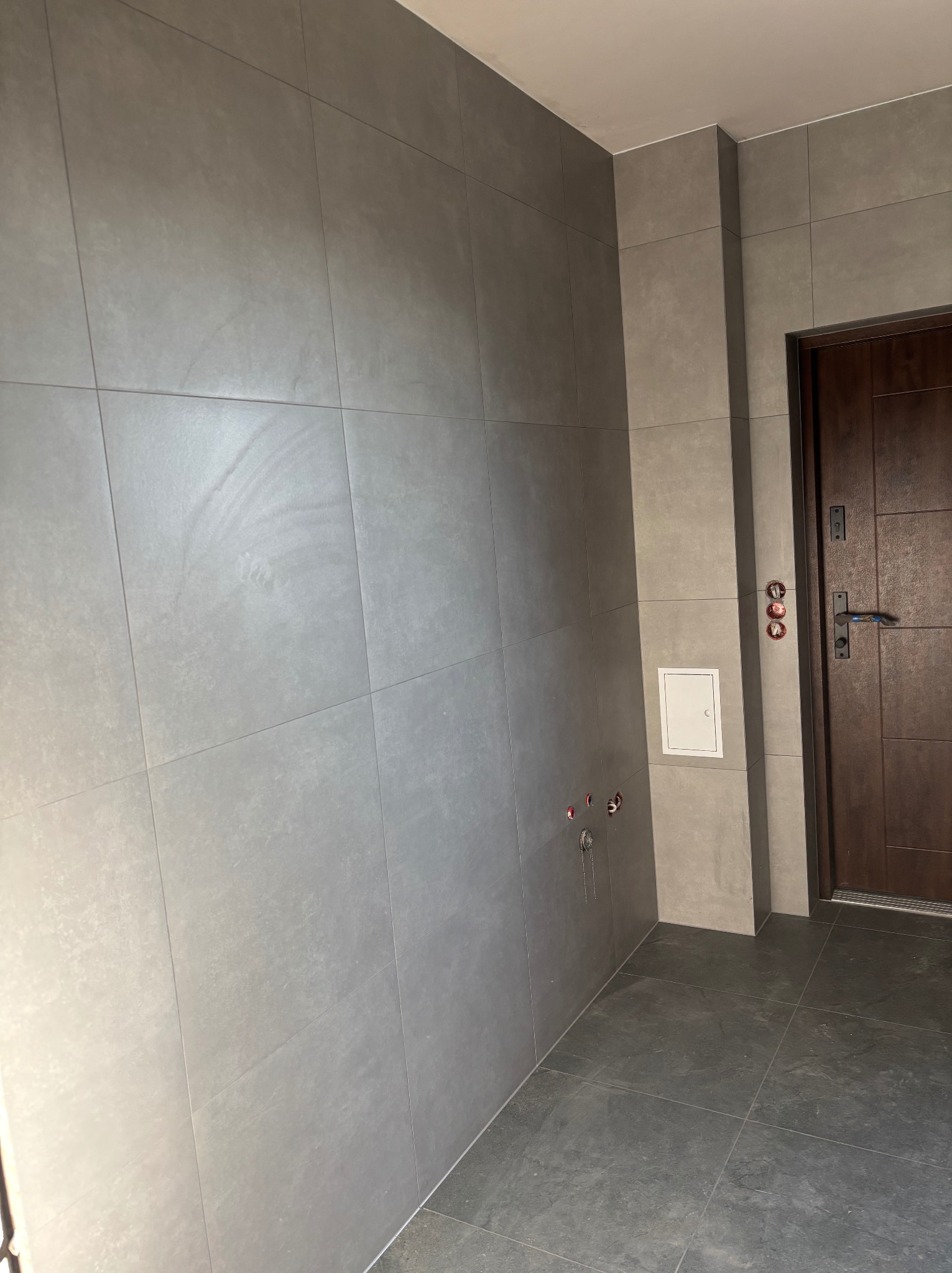 Szara, matowa okładzina ścienna z płytek w korytarzu, z widoczną brązową stolarką drzwiową i elementami instalacji wodnej. Minimalistyczny design.