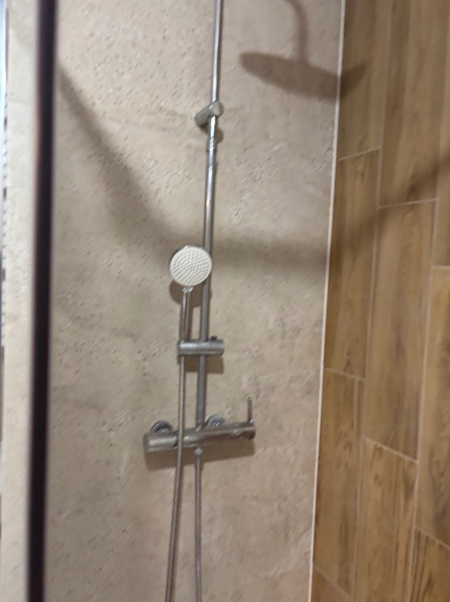 Nowoczesny zestaw prysznicowy z deszczownicą na tle beżowych płytek imitujących kamień i drewnopodobnych płytek. Widoczne chromowane elementy.