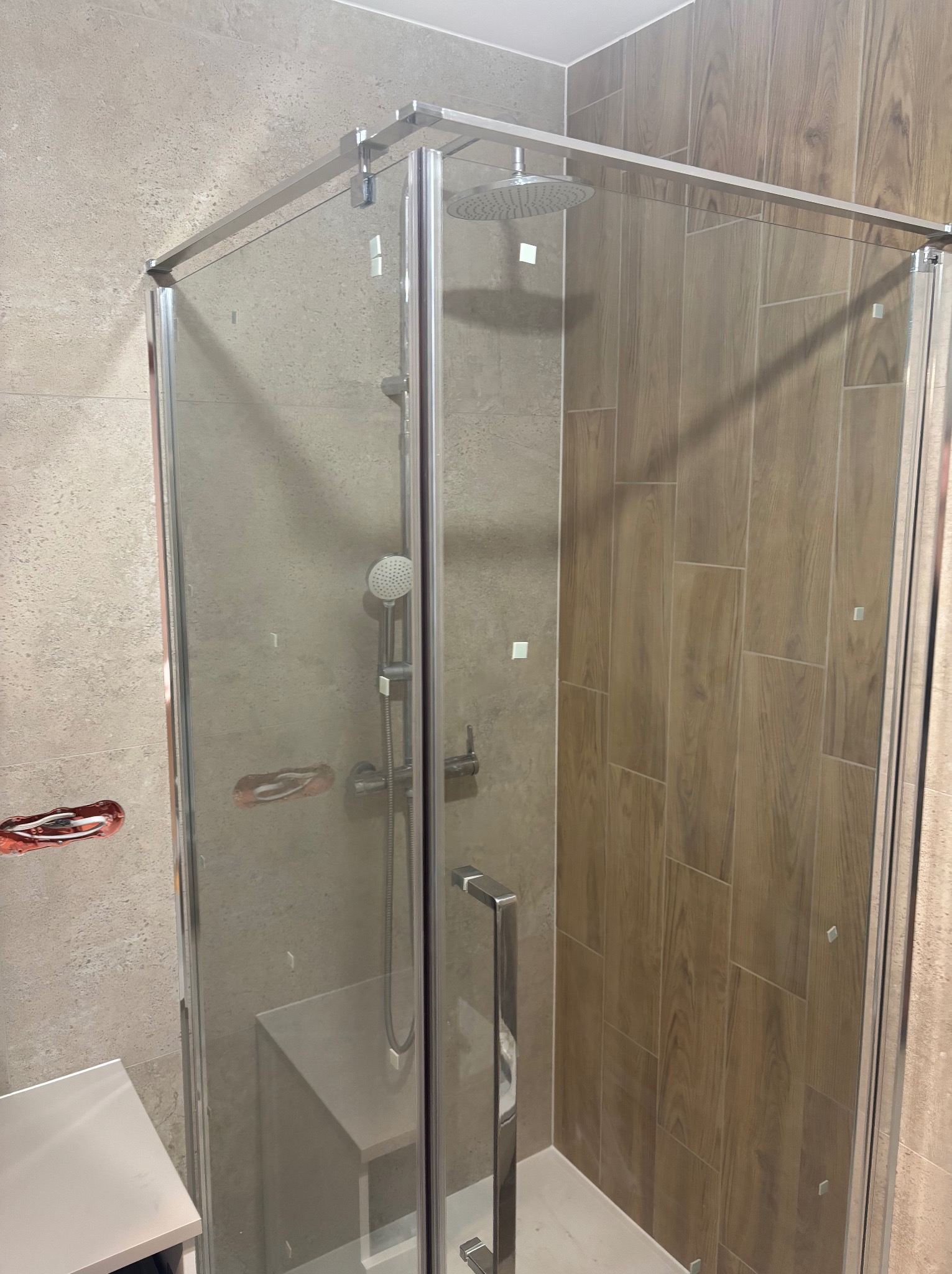 Nowoczesna kabina prysznicowa z chromowanymi profilami i deszczownicą, obłożona płytkami imitującymi drewno, z widoczną instalacją wodną w ścianie.