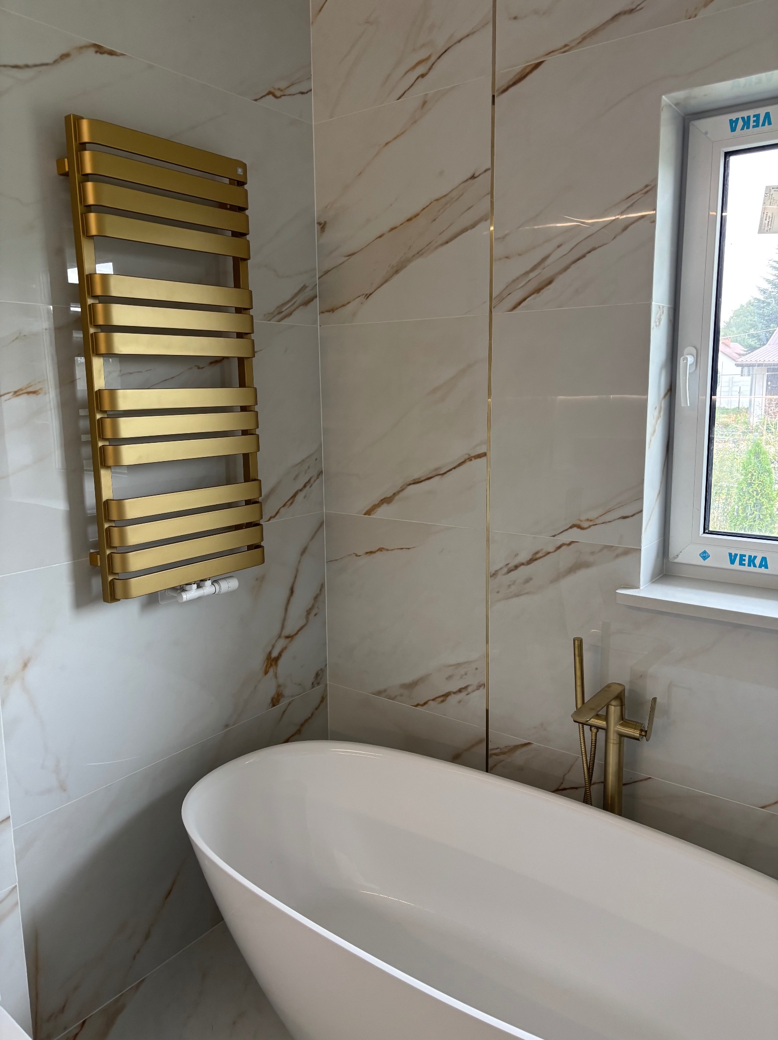 Elegancka łazienka z wanną wolnostojącą, złotą armaturą i grzejnikiem drabinkowym na tle marmurowych płytek. Widok na okno z widokiem na zieleń.