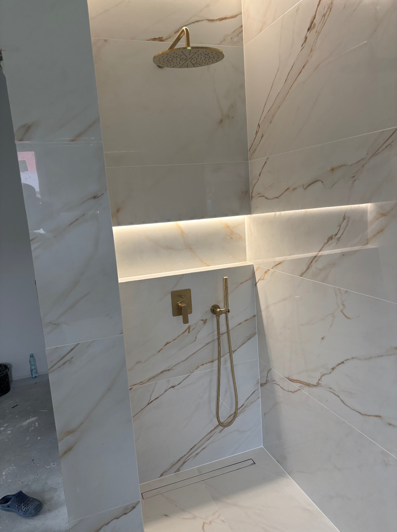 Wykończona łazienka z marmurowymi płytkami w odcieniach bieli i złota, wyposażona w złotą armaturę i podświetlaną wnękę. Widoczny odpływ liniowy.