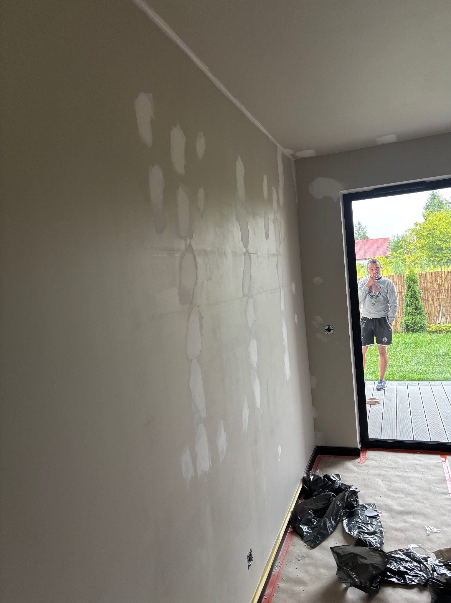 Ściana w trakcie szpachlowania z widocznymi łatami gładzi, zabezpieczona podłoga folią i osoba w tle, stojąca w otwartych drzwiach na zewnątrz.