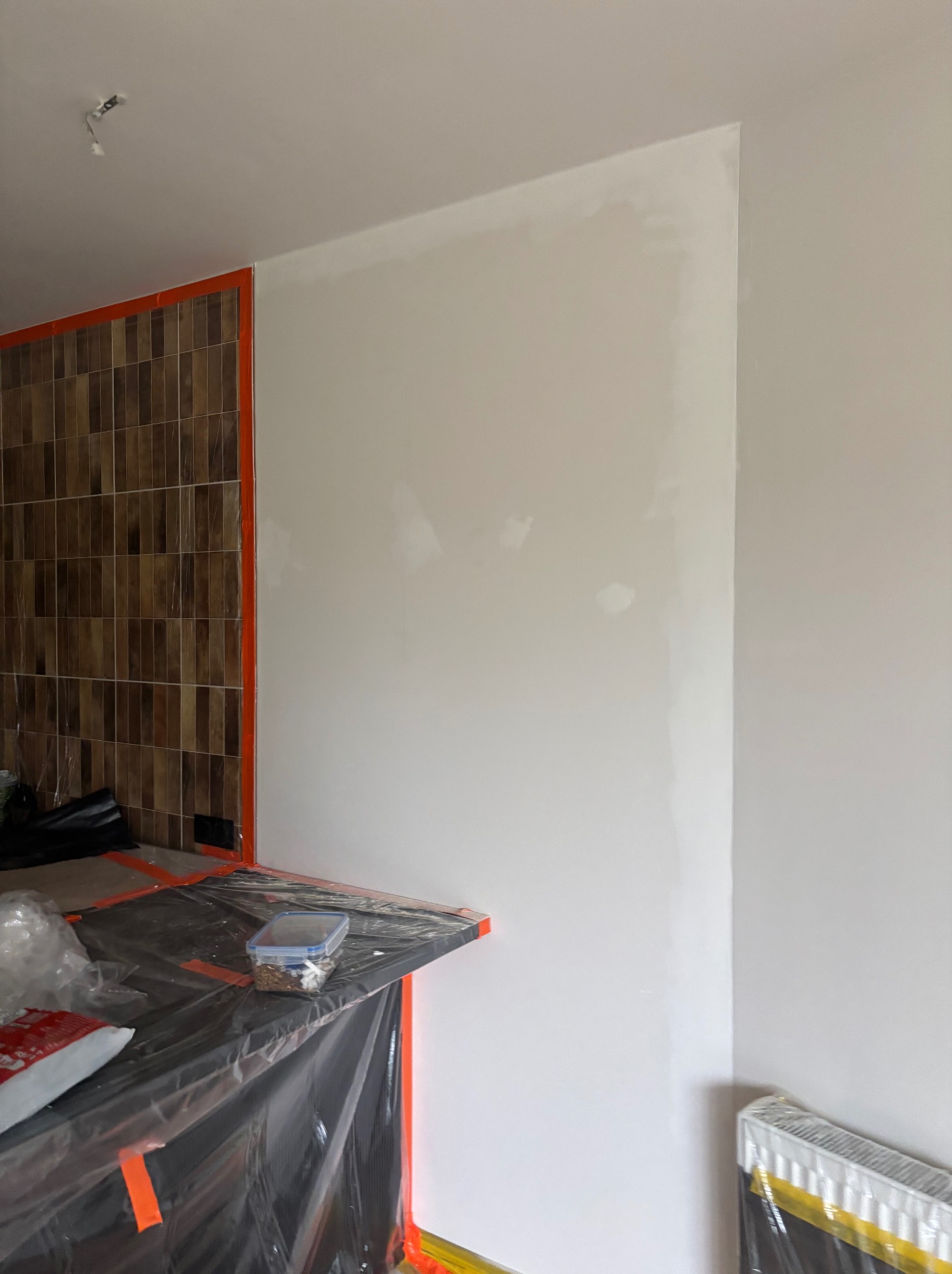 Ściana po szpachlowaniu, przygotowana do malowania. Widoczne zabezpieczenia folią malarską i taśmą. Fragment ściany z brązowymi płytkami.