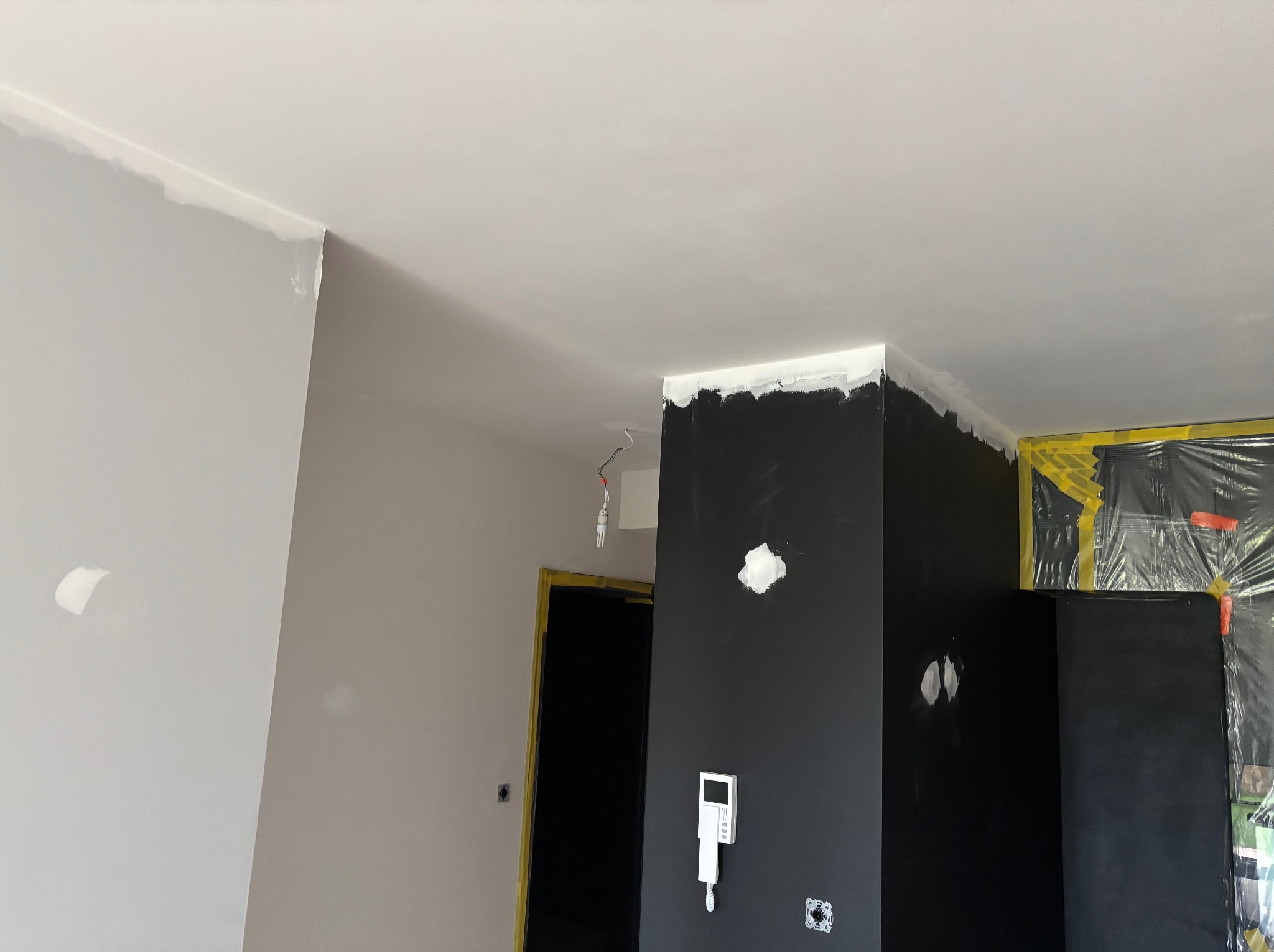 Wnętrze w trakcie malowania: czarna ściana z widocznymi śladami szpachlowania i świeżo pomalowany sufit na biało. Zabezpieczone folią drzwi i fragment ściany w odcieniach szarości.
