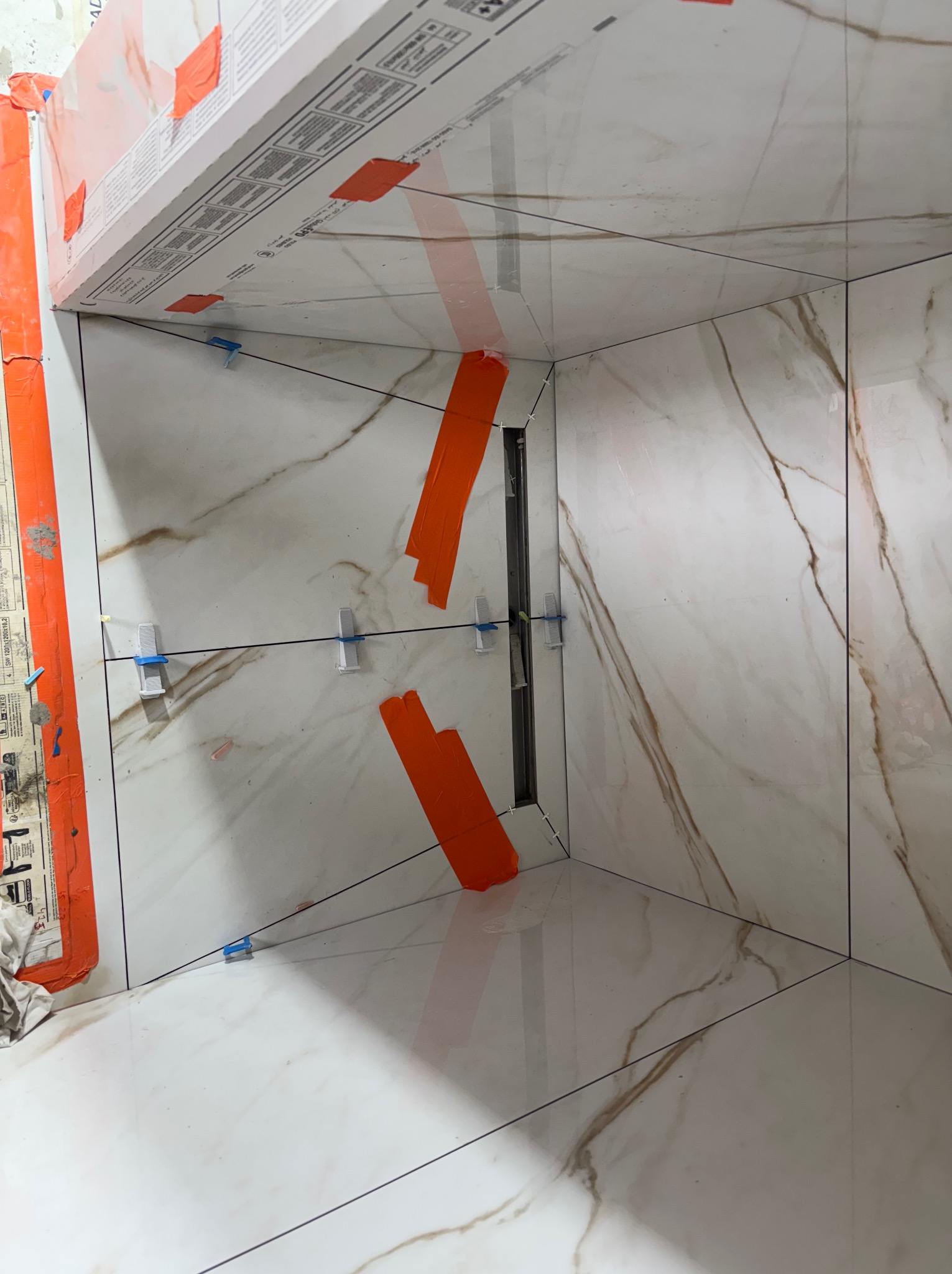 Wnętrze kabiny prysznicowej w trakcie prac glazurniczych. Widoczne białe płytki z marmurkowym wzorem, pomarańczowa taśma, niebieskie krzyżyki dystansowe i odpływ liniowy.