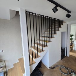 Dariusz Ernest PRECYZJA SPAWANIA - Nowoczesne schody z drewnianymi stopniami i czarną metalową balustradą w jasnym wnętrzu. Widoczne narzędzia sugerują prace wykończeniowe.