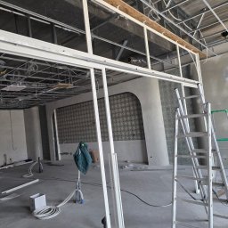 Dariusz Ernest PRECYZJA SPAWANIA - Montaż stalowej konstrukcji ścian działowych w surowym wnętrzu. Widoczne profile, drabiny i instalacje pod sufitem. Etap prac wykończeniowych.