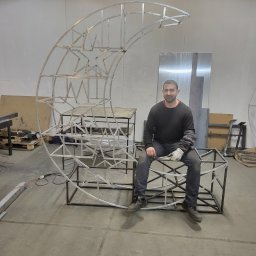 Dariusz Ernest PRECYZJA SPAWANIA - Spawacz siedzi na konstrukcji metalowej w kształcie księżyca i gwiazd. Widoczne spawy i elementy warsztatu, co podkreśla rzemieślniczy charakter usługi.