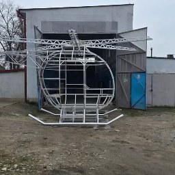 Dariusz Ernest PRECYZJA SPAWANIA - Szkielet helikoptera ze spawanych rur, ustawiony przed budynkiem warsztatowym z otwartymi wrotami. Widoczny fragment podwozia, wirnik ogonowy i główna rama.