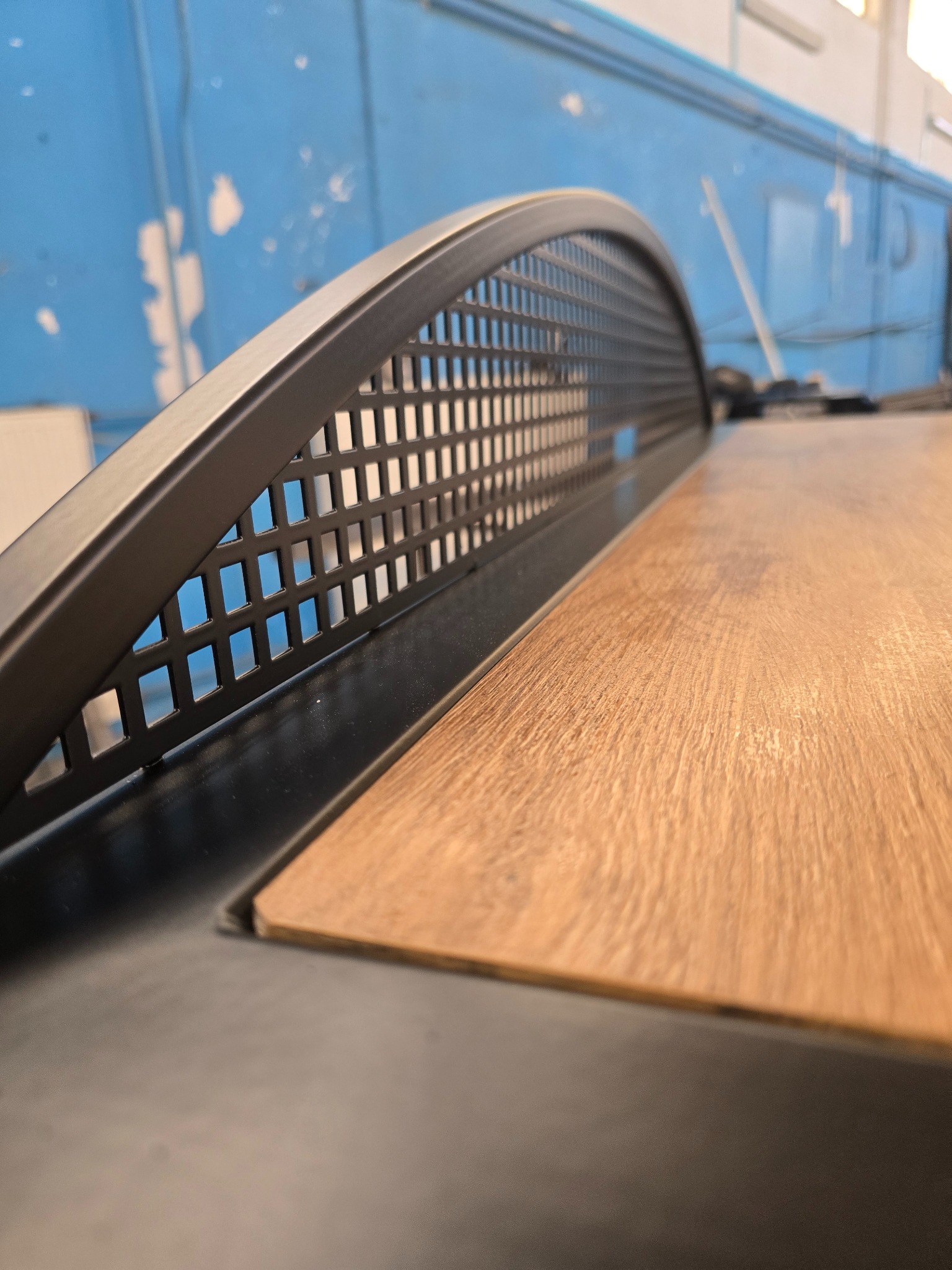 Metalowa osłona z perforacją i drewniany blat w zbliżeniu, ukazujące precyzję wykonania detali konstrukcji. Niebieskie tło z przetarciami dodaje charakteru.