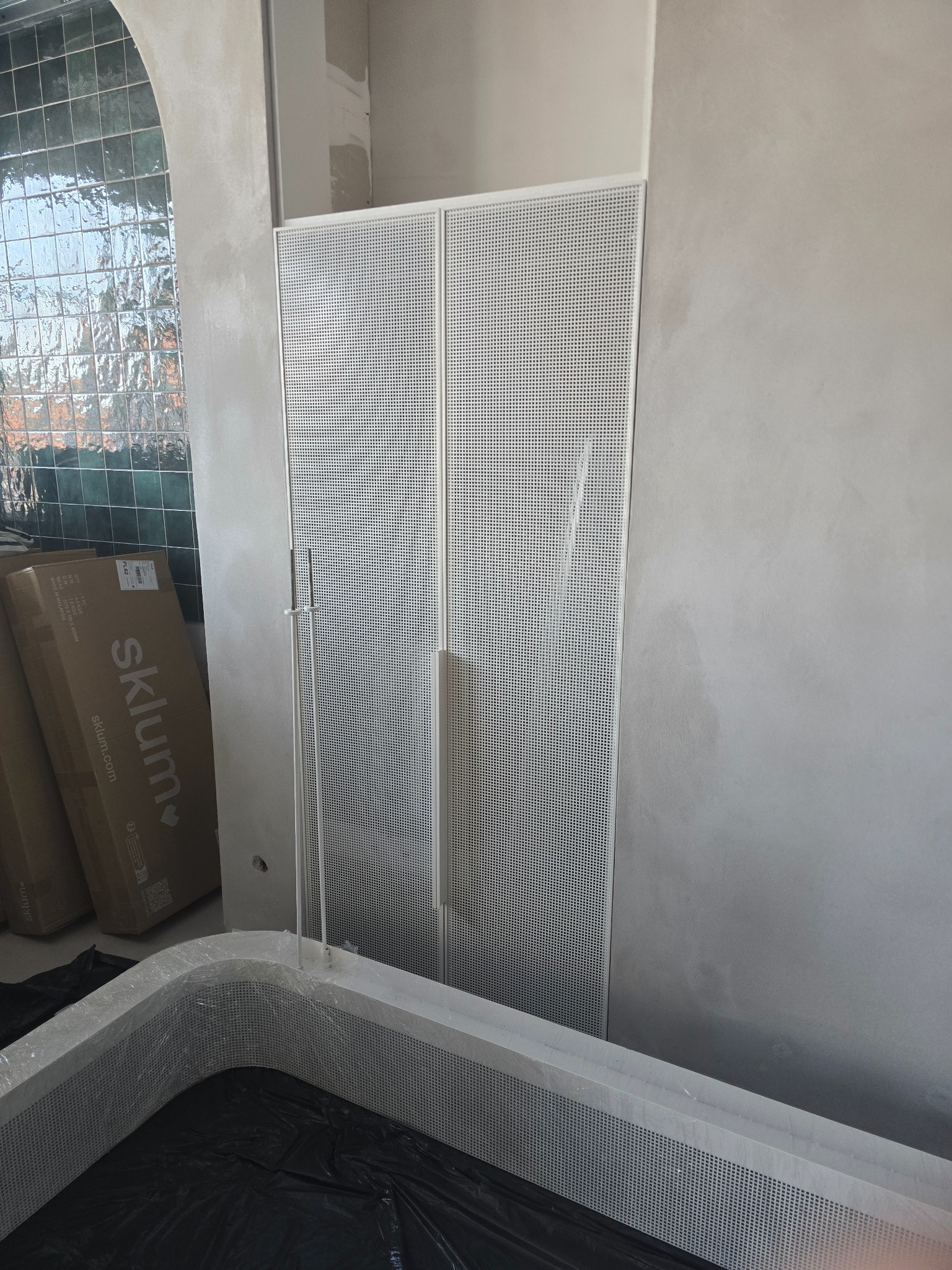 Spawana, perforowana konstrukcja obudowy w łazience, z widocznymi elementami wykończeniowymi i paczkami w tle. Metalowa obudowa w trakcie montażu.