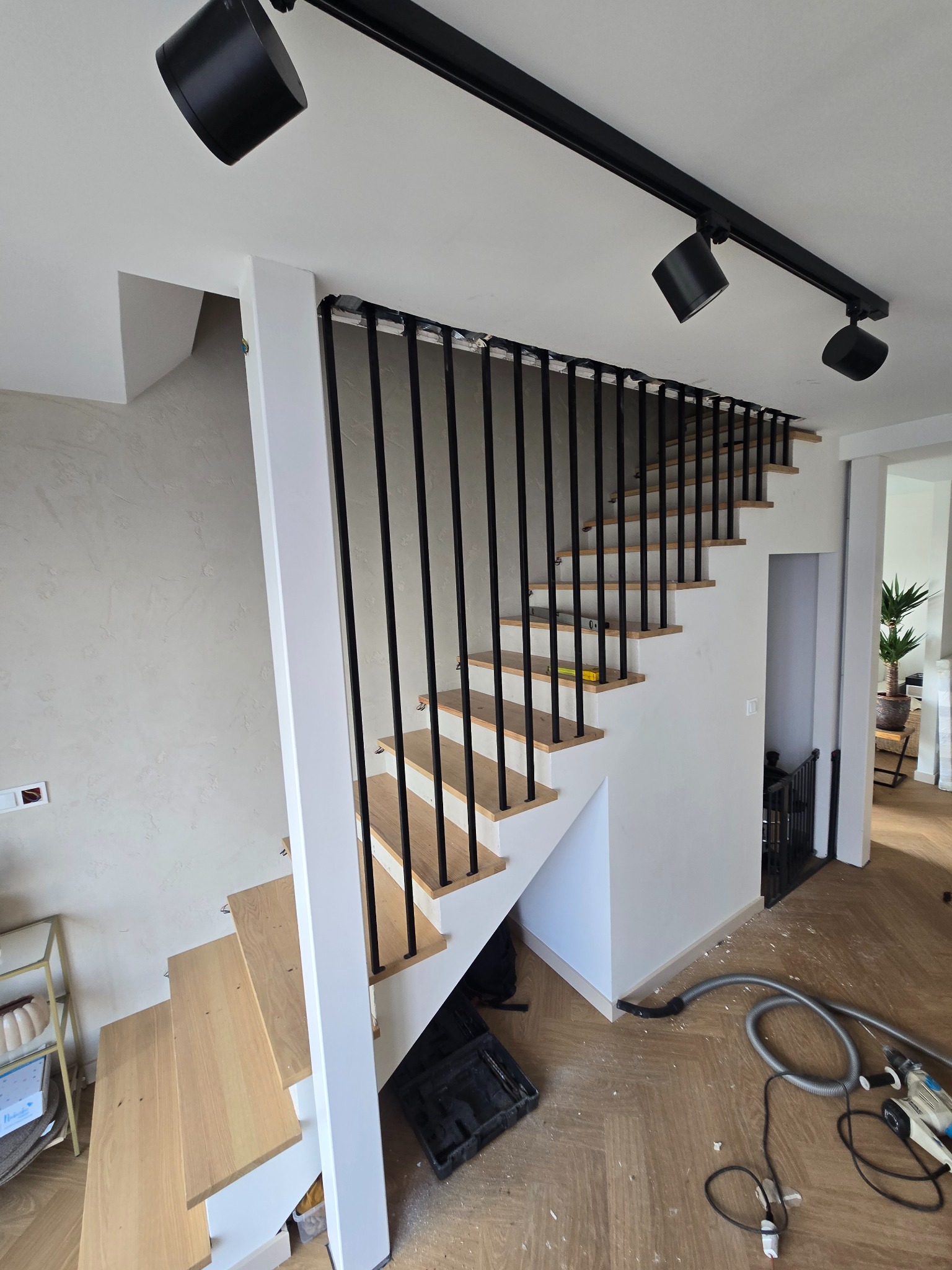 Nowoczesne schody z drewnianymi stopniami i czarną metalową balustradą w jasnym wnętrzu. Widoczne narzędzia sugerują prace wykończeniowe.