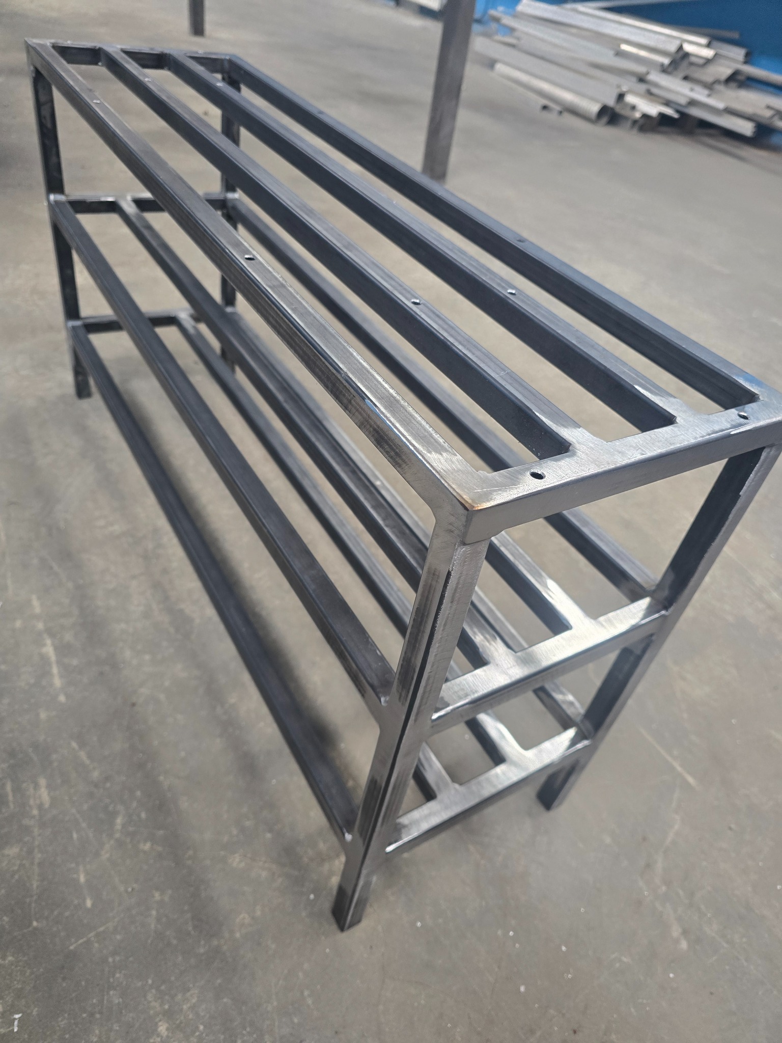 Ręcznie wykonany, metalowy regał na buty z trzema poziomami, spawany z profili o kwadratowym przekroju, surowy metal, widoczne spawy.