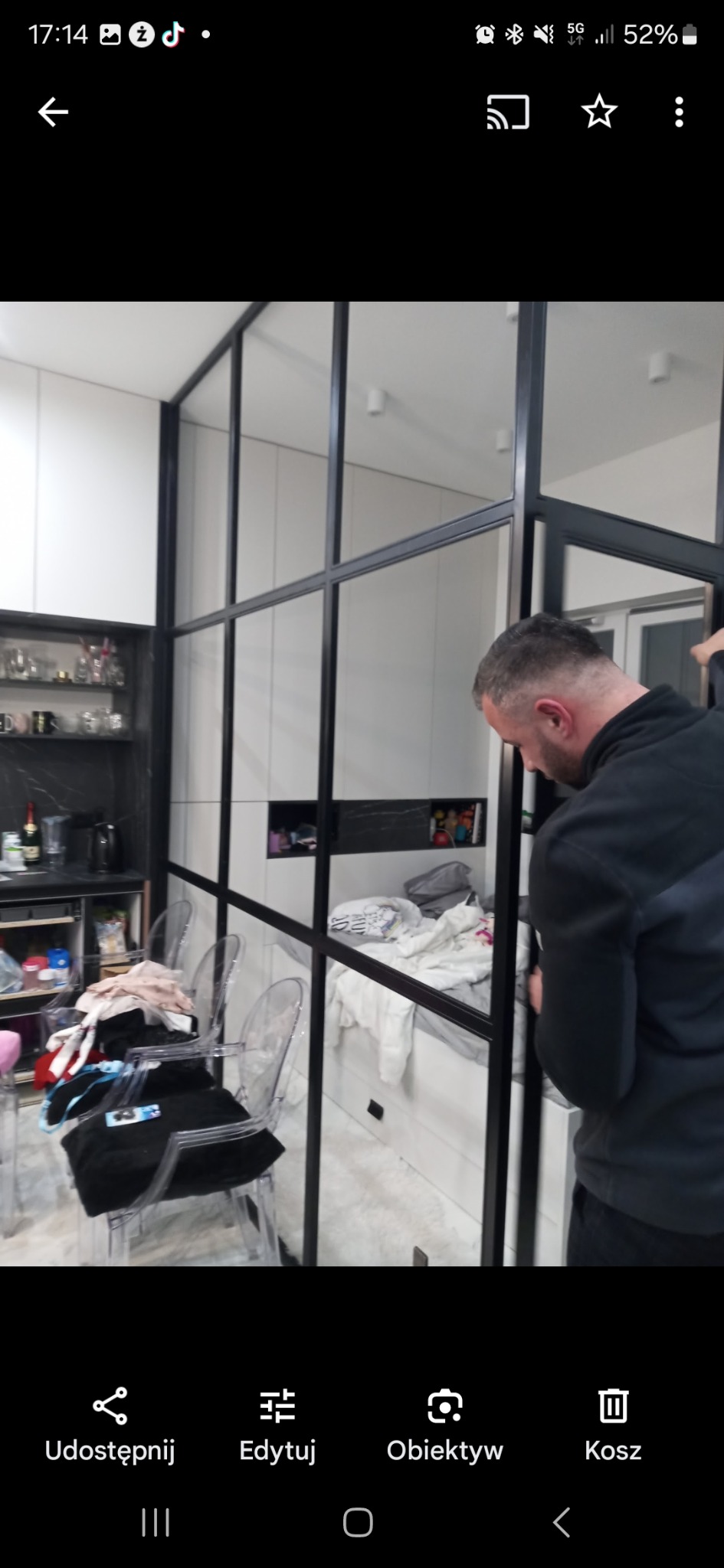 Montaż industrialnej ścianki działowej z czarnych profili w nowoczesnym wnętrzu. Widoczna częściowo osoba montująca konstrukcję, transparentne krzesła i wyposażenie kuchni w tle.