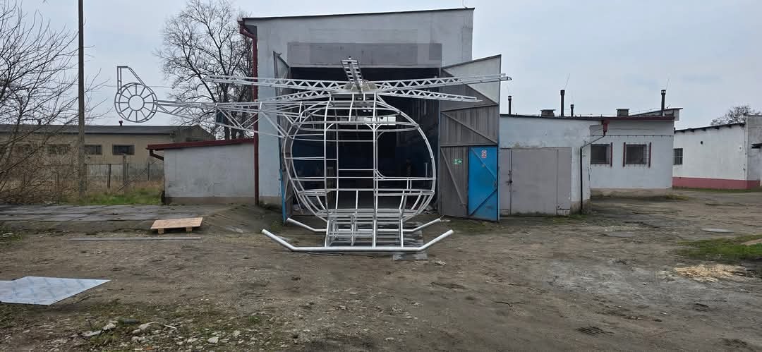 Szkielet helikoptera ze spawanych rur, ustawiony przed budynkiem warsztatowym z otwartymi wrotami. Widoczny fragment podwozia, wirnik ogonowy i główna rama.