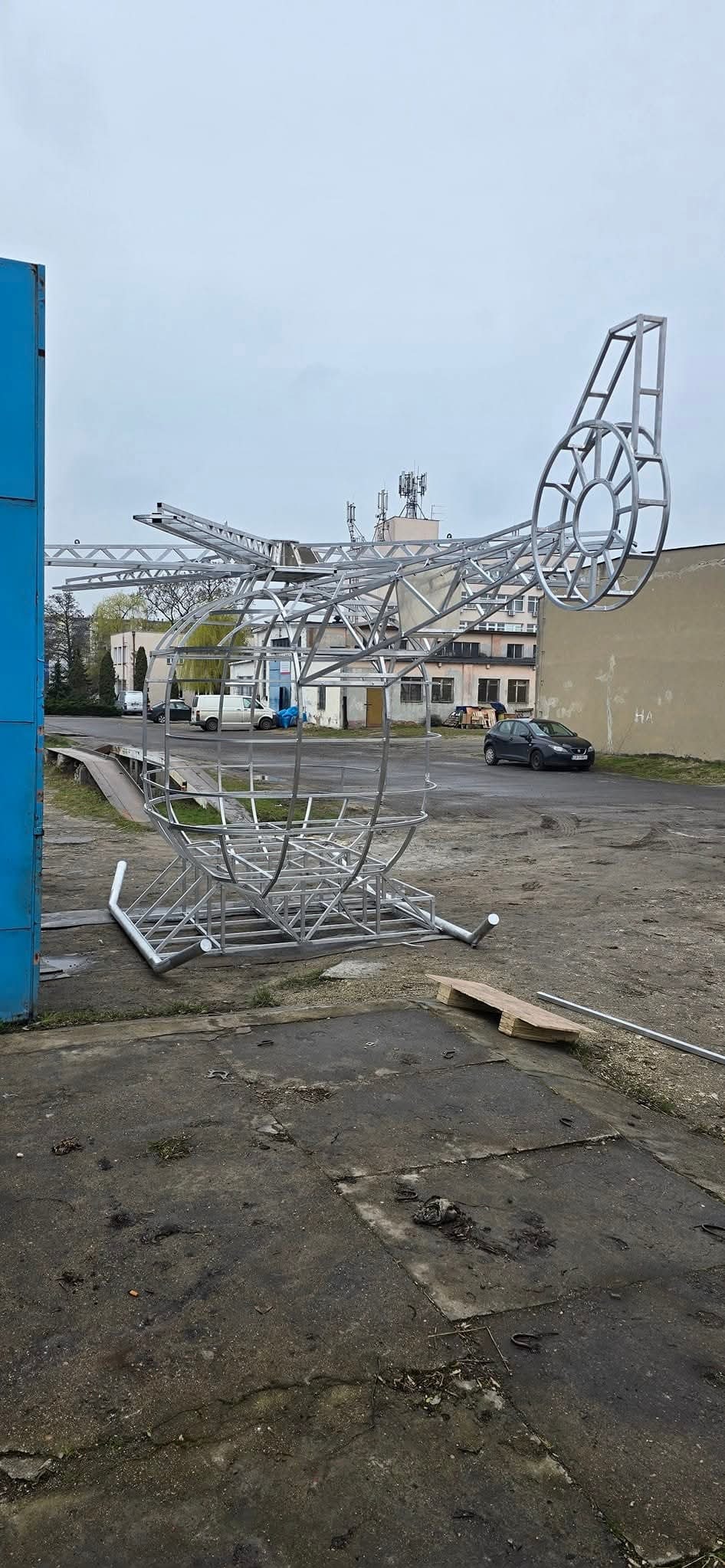 Metalowa konstrukcja szkieletu helikoptera na zewnątrz, z detalami spawów i elementami montażowymi. W tle budynki i samochód.