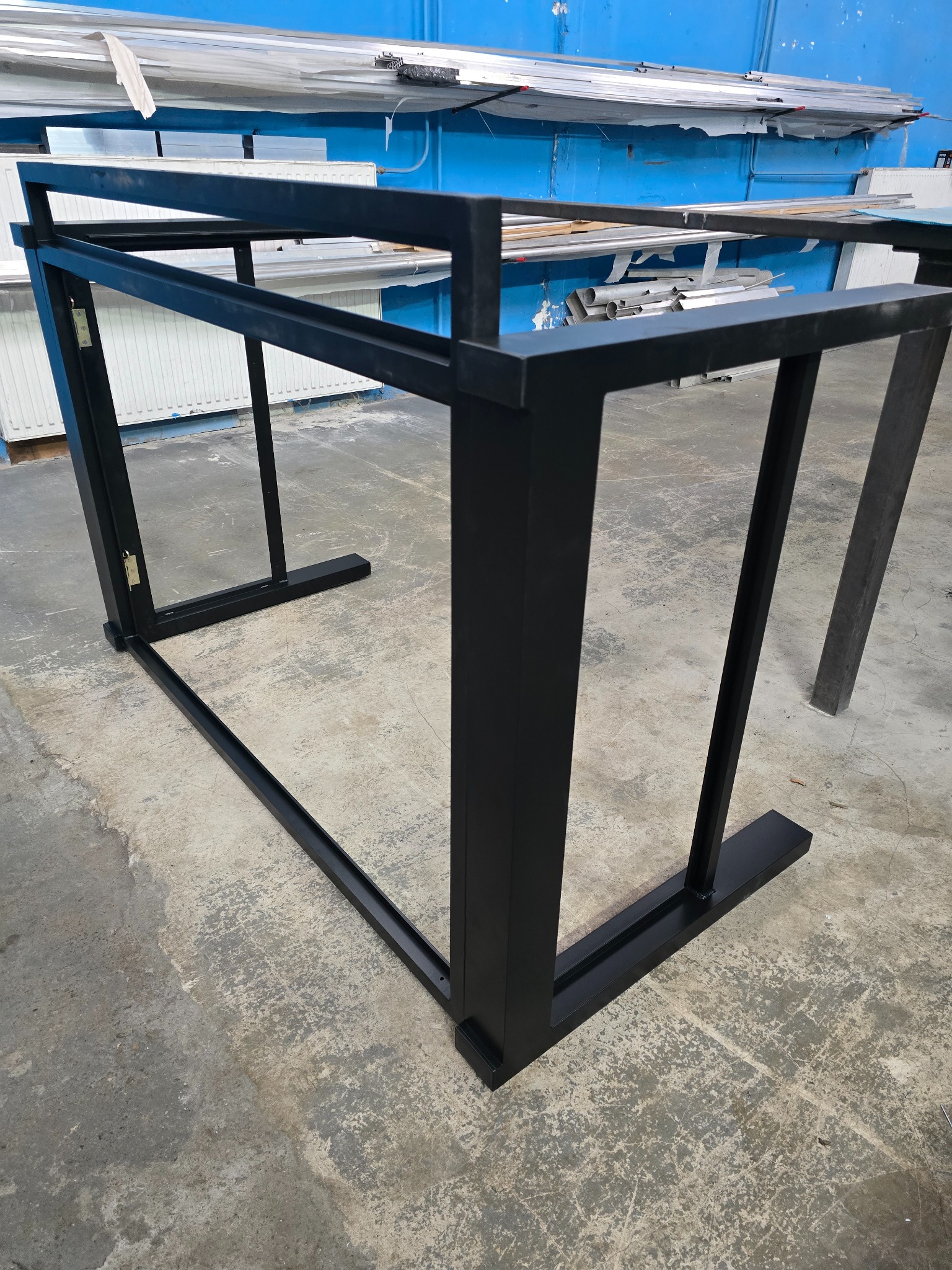 Czarna, spawana konstrukcja stalowa w warsztacie, rama z profili zamkniętych, widoczne spawy i zawiasy, surowy beton na podłodze, niebieska ściana w tle.