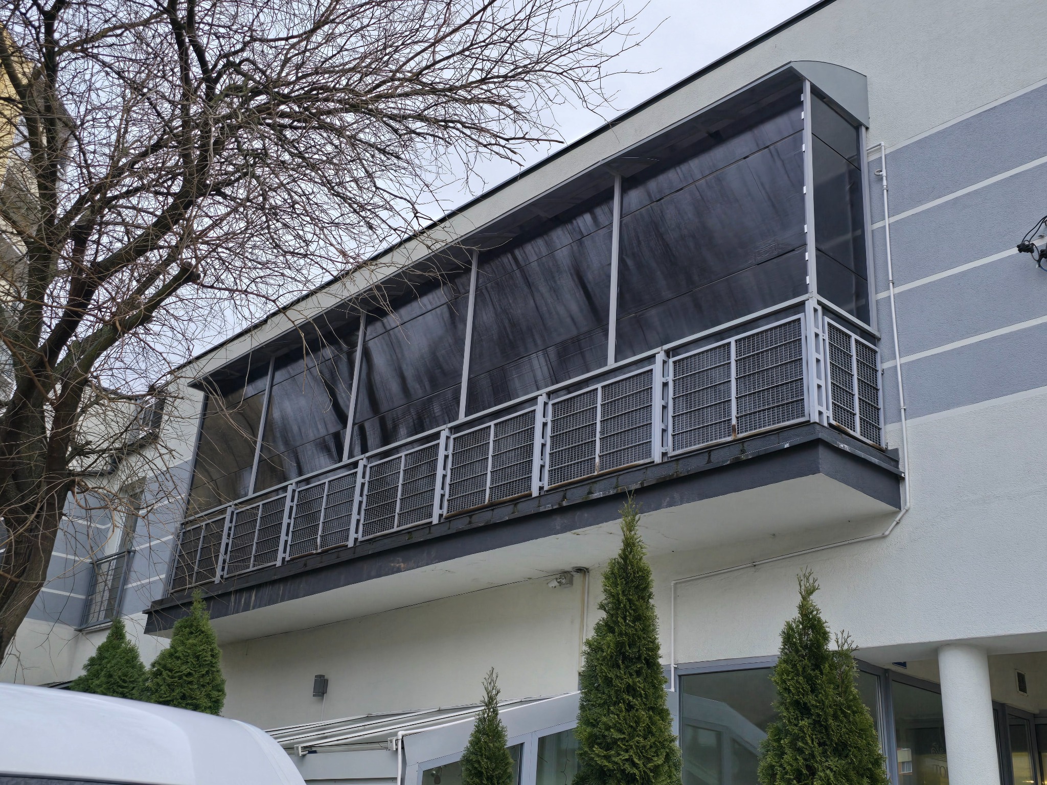 Zewnętrzny balkon z metalową kratą i czarną osłoną, widoczny z dołu. W tle drzewo bez liści i fragment białego samochodu. Szare tło budynku z kolumnami.