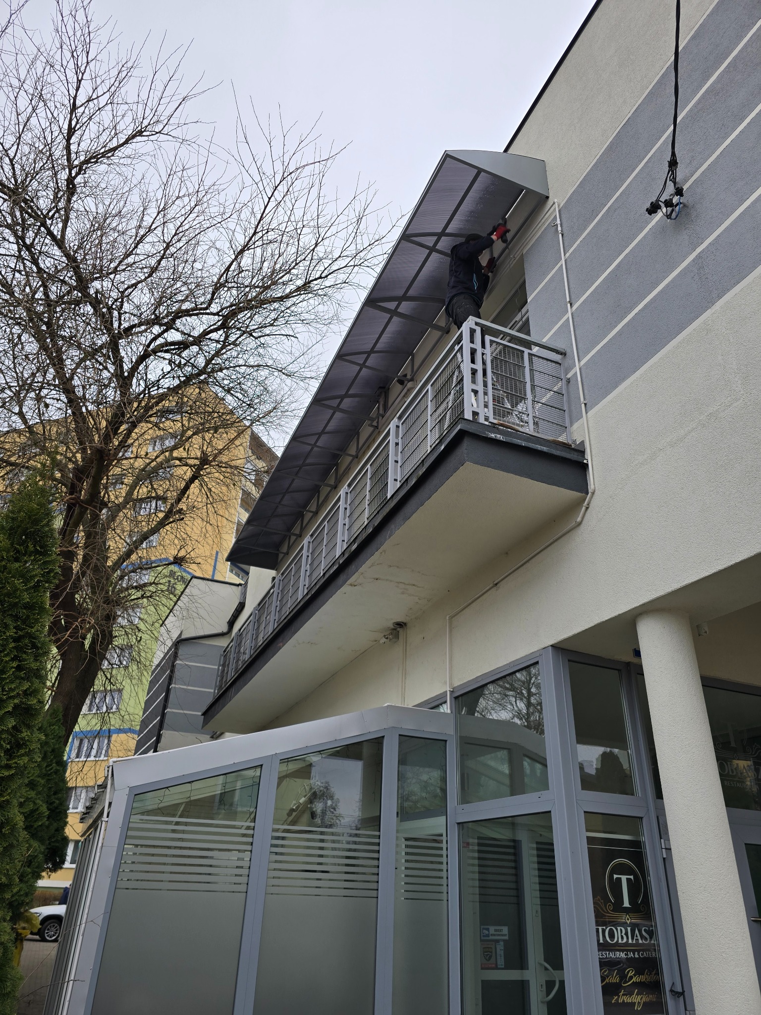Montaż zadaszenia nad balkonem budynku z szarymi pasami. Widoczna osoba w trakcie pracy, drzewo bez liści i fragment budynku z witryną restauracji.