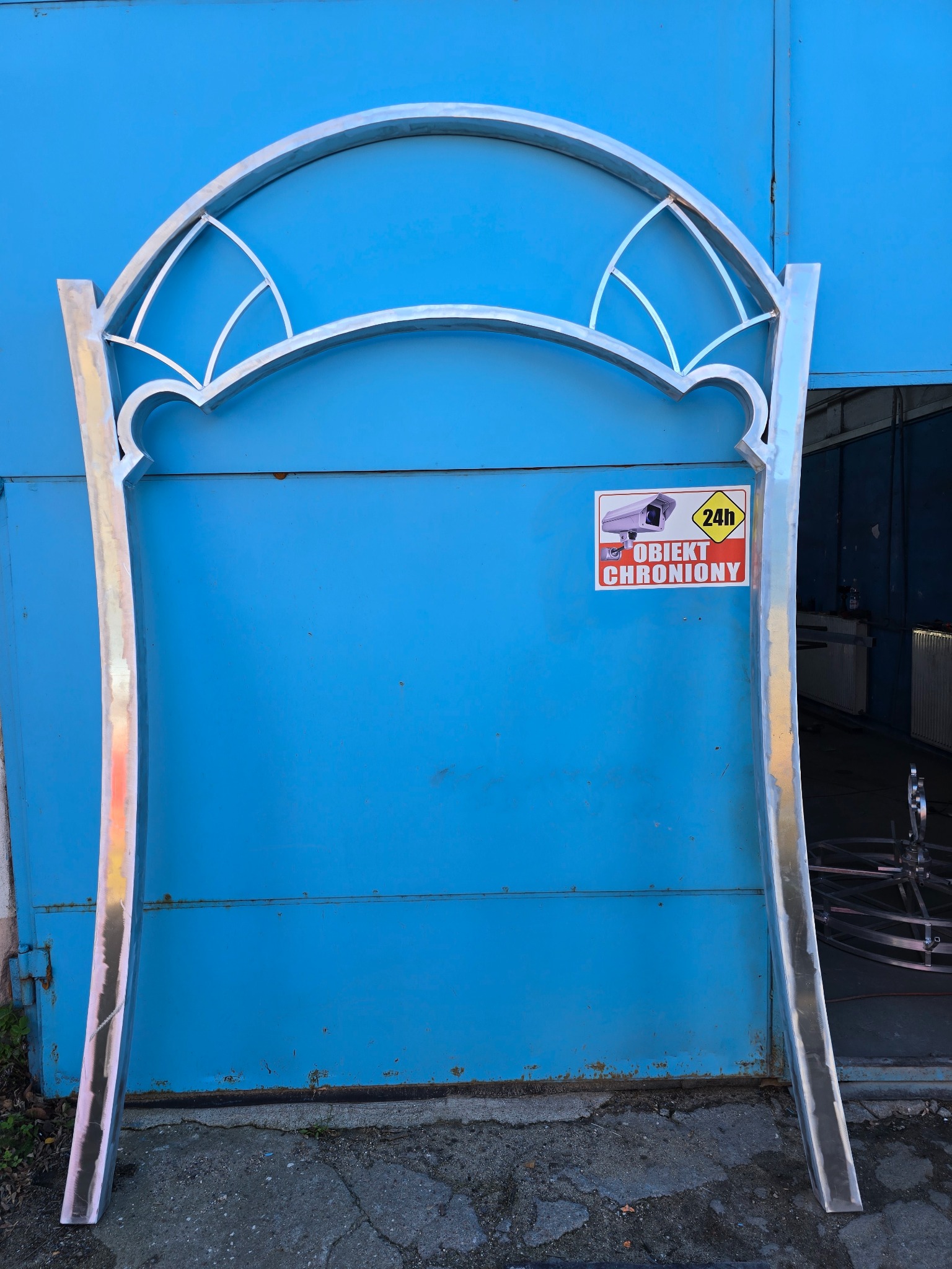 Metalowa konstrukcja łukowa z dekoracyjnym wzorem, prawdopodobnie element bramy lub ogrodzenia, na tle niebieskich drzwi garażowych z naklejką 'Obiekt Chroniony'.