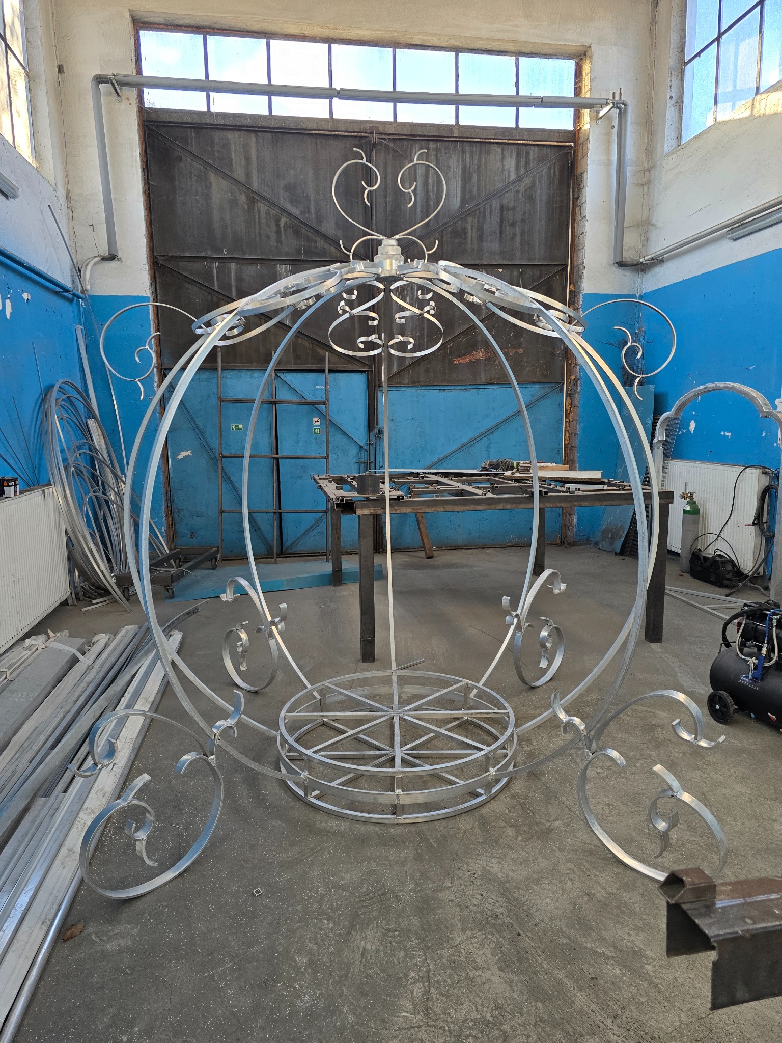 Metalowa konstrukcja powozu w warsztacie, spawana z profili, widoczne elementy dekoracyjne i narzędzia. Niebieskie ściany, stalowe drzwi w tle.