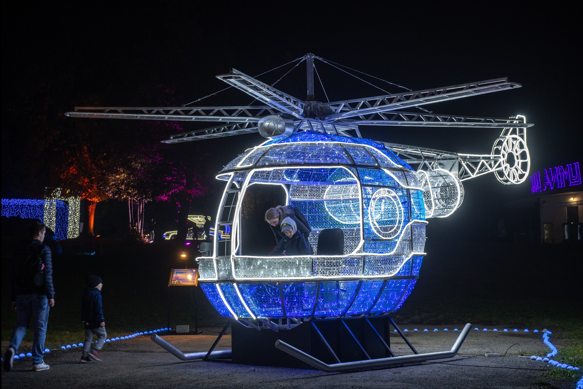Iluminowany helikopter z metalową konstrukcją, wewnątrz dwie osoby. Dekoracja świetlna w parku, w tle drzewa i budynek z neonem.