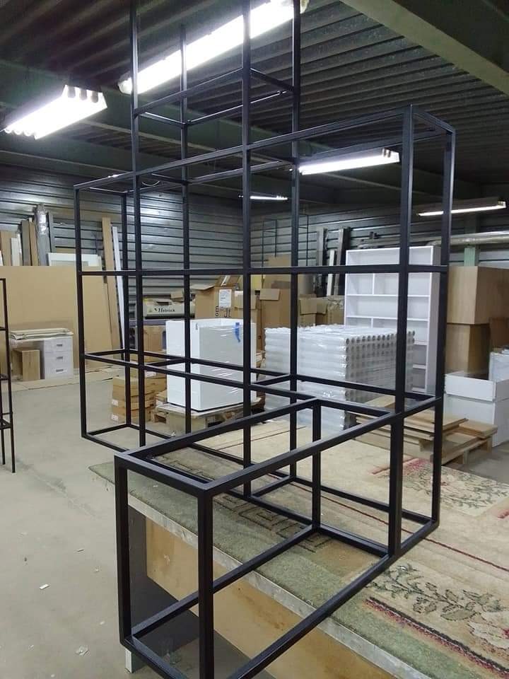Metalowa konstrukcja regału z kwadratowych profili, czarne wykończenie, widoczne spawy, industrialny styl, w tle magazyn z kartonami i meblami.