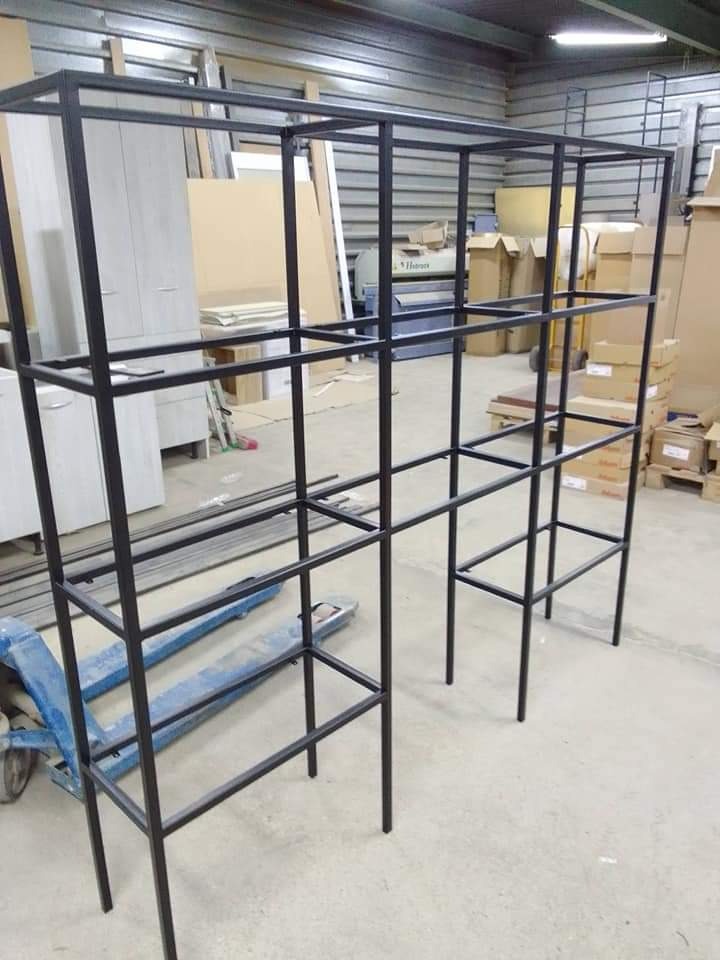 Czarna, spawana konstrukcja regału z metalowych profili, ustawiona w warsztacie na tle kartonów i maszyny do obróbki metali. Widoczny wózek paletowy.