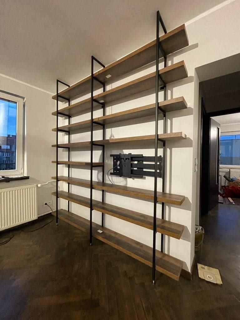 Regał z drewnianymi półkami i metalową konstrukcją, zamontowany na ścianie, z uchwytem na telewizor. Widok na parkiet i okno w tle.